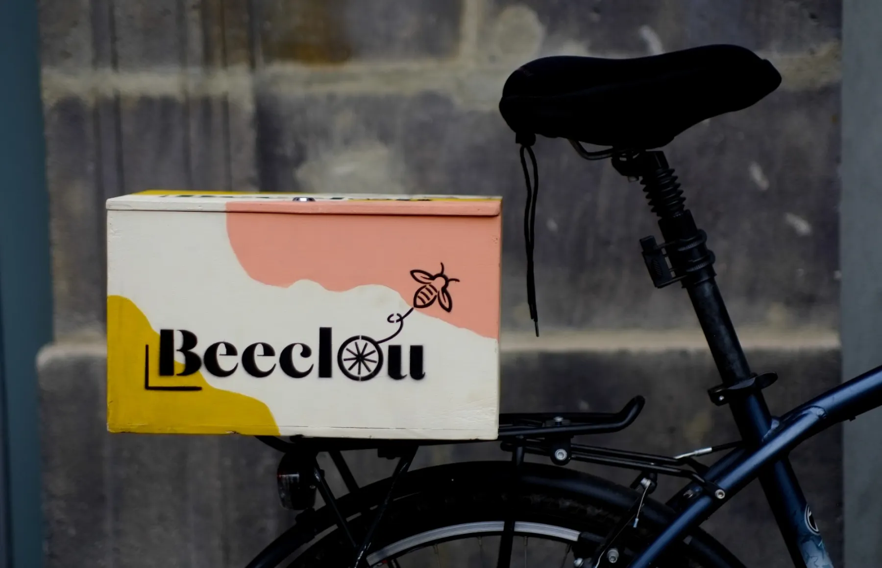 La boîte Beeclou jaune, crème et rose sur un porte-bagages, devant un mur de pierre.